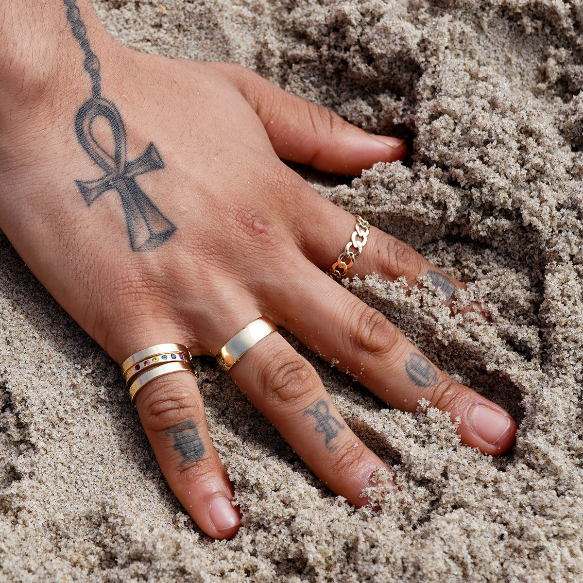 A model with their hand in the sand at the beach wearing various Automic Gold rings including a Curb Ring