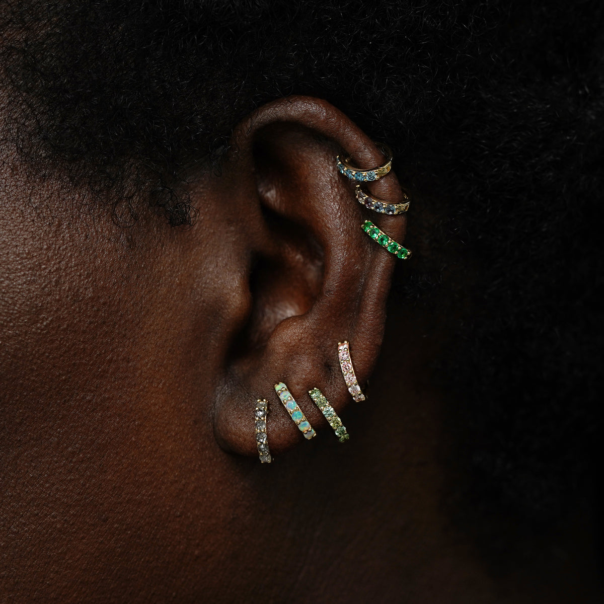 A small gemstone huggie hoop above a mini gemstone huggie hoop on a model's finger to show size difference