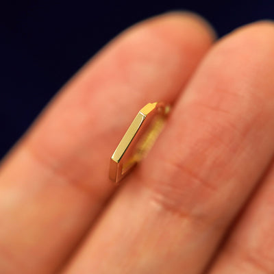 A 14k solid gold Hexagon Huggie Hoop between a model's fingers to show the thickness of the earring