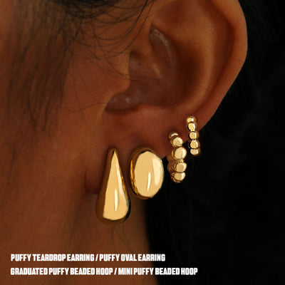 A yellow gold Oval Puffy Earring, Teardrop Puffy Earring, Mini Puffy Beaded Hoop, and Graduated Puffy Beaded Hoop on one ear