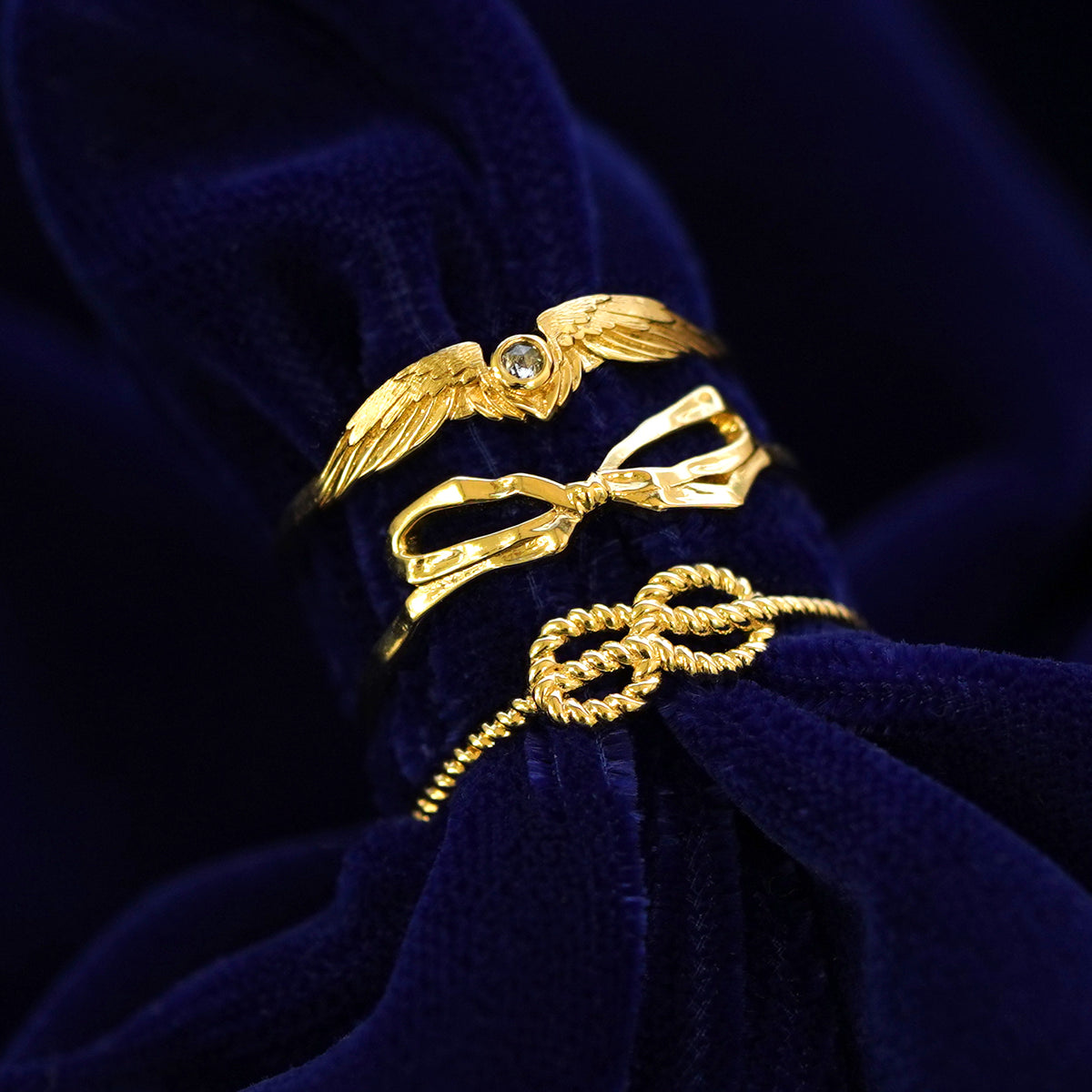 A Diamond Wings Ring, Bow Ring, and Infinity Rope Ring stacked on a dark blue cloth