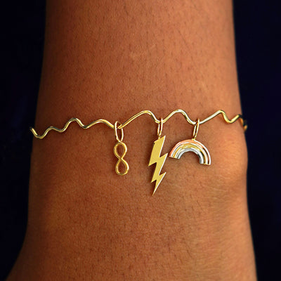 A model's wrist wearing a yellow gold Wave Bangle with an Infinity Charm, a Lightning Bolt Charm, and a Rainbow Charm