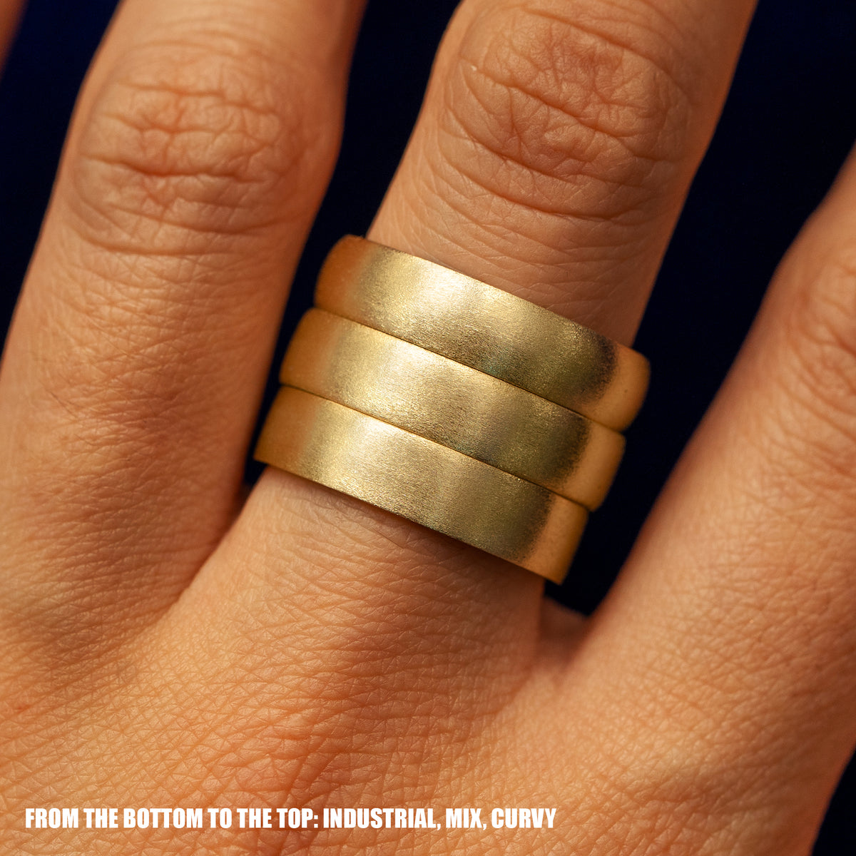 Close up view of a model's fingers wearing an Industrial Band Mix Band and Curvy band all in yellow gold 