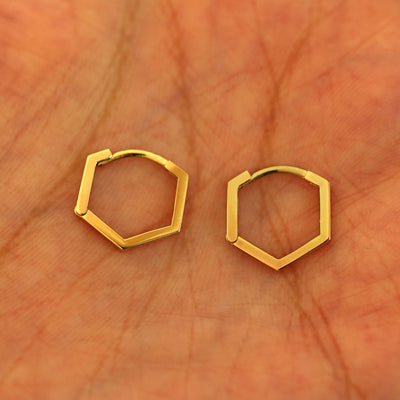 A pair of 14k yellow gold Hexagon Huggie Hoops resting in a model's palm