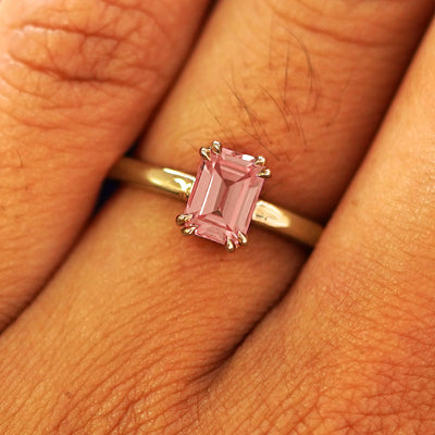 Close up view of a model's fingers wearing a 14k yellow gold Emerald Cut Lab Pink Sapphire Ring