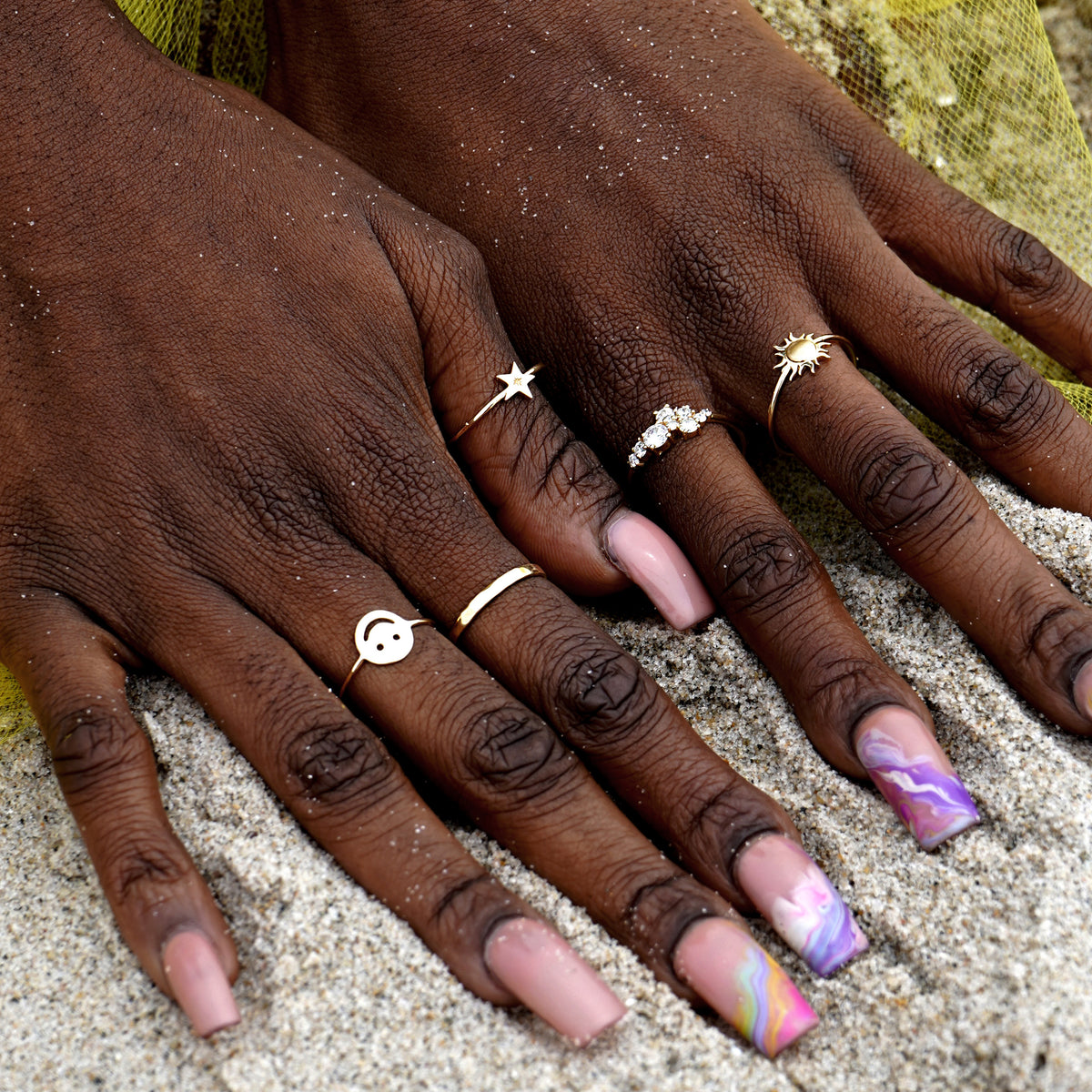 A model's hands with long nails digging into sand at the beach wearing a Diamond Cluster ring and other Automic Gold rings